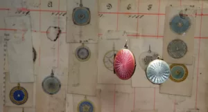 Rectangular bookpage featuring coloured drawings of round pendants. An red oval and a round light blue locket pendant are placed in front of the page hanging almost invisibily from colourless wire strings.