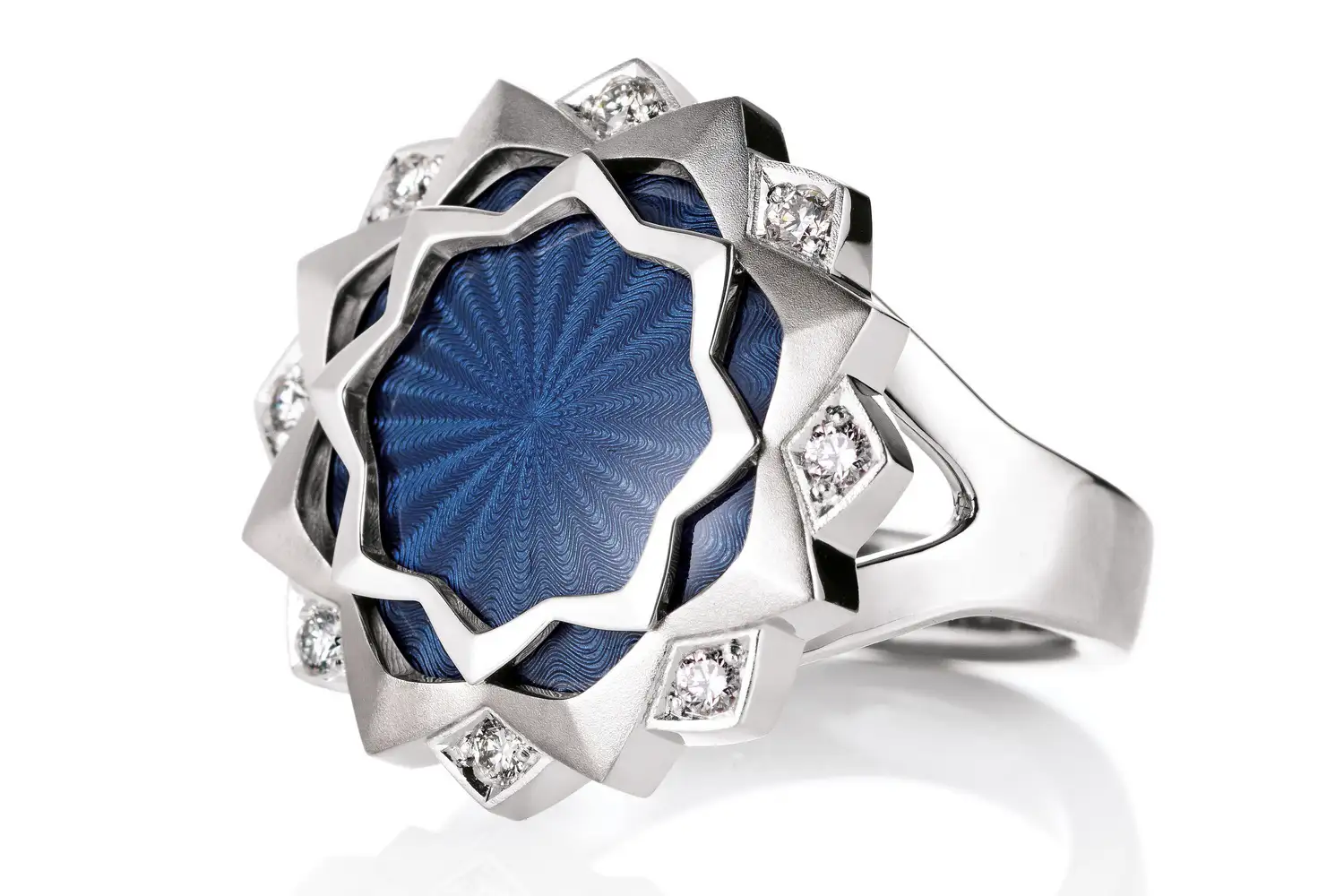 Ring Chrysanthemum: 18kt white gold, indigo blue vitreous enamel, guilloche engraving by hand, 8 diamonds, total 0.20 ct, G VS, diameter of ring head 21,5 mm. Limited edition of 50 pieces. V1696-HB-00-00-105,1500×1000. Horizontal picture. Silver-coloured, round ring with star-shaped decoration and blue, transparent colour, under which fine wave patterns are visible. Set all round with 8 white diamonds,1500x1000. Horizontal picture.