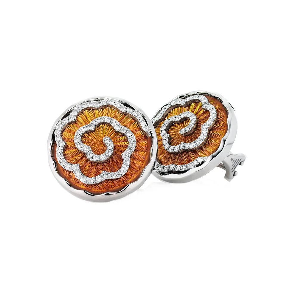 Ear clips made of white gold with a round head. The head is about the size of a large coin. Its surface shimmers in a strong golden-amber tone with a wave-like pattern. On top lies the shape of a large flower set with many small diamonds.