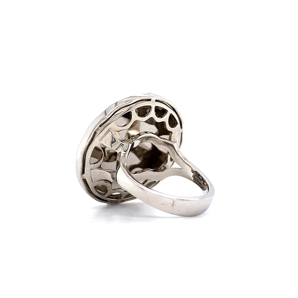 A ring made of white gold with a round head. The head is about the size of a large coin. Its surface shimmers in a strong golden-amber tone with a wave-like pattern. On top lies the shape of a large flower set with many small diamonds.