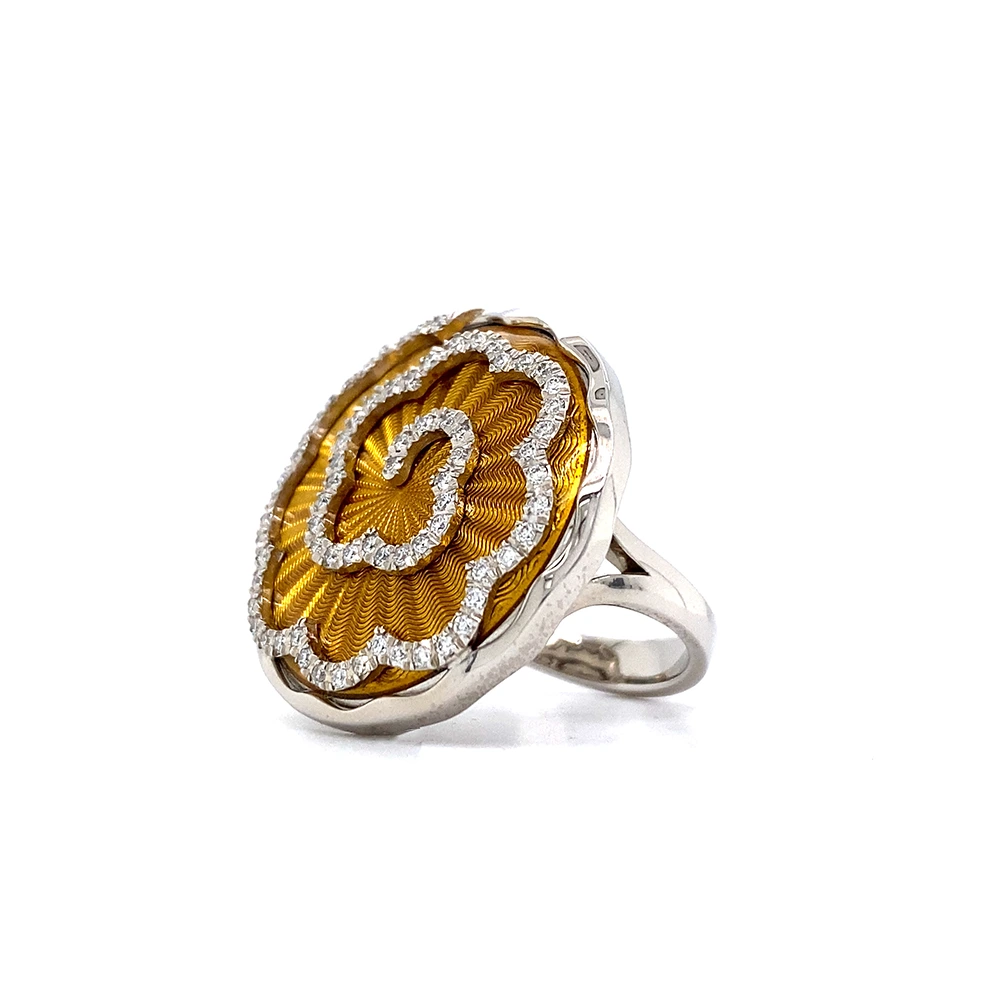 A ring made of white gold with a round head. The head is about the size of a large coin. Its surface shimmers in a strong golden-amber tone with a wave-like pattern. On top lies the shape of a large flower set with many small diamonds.