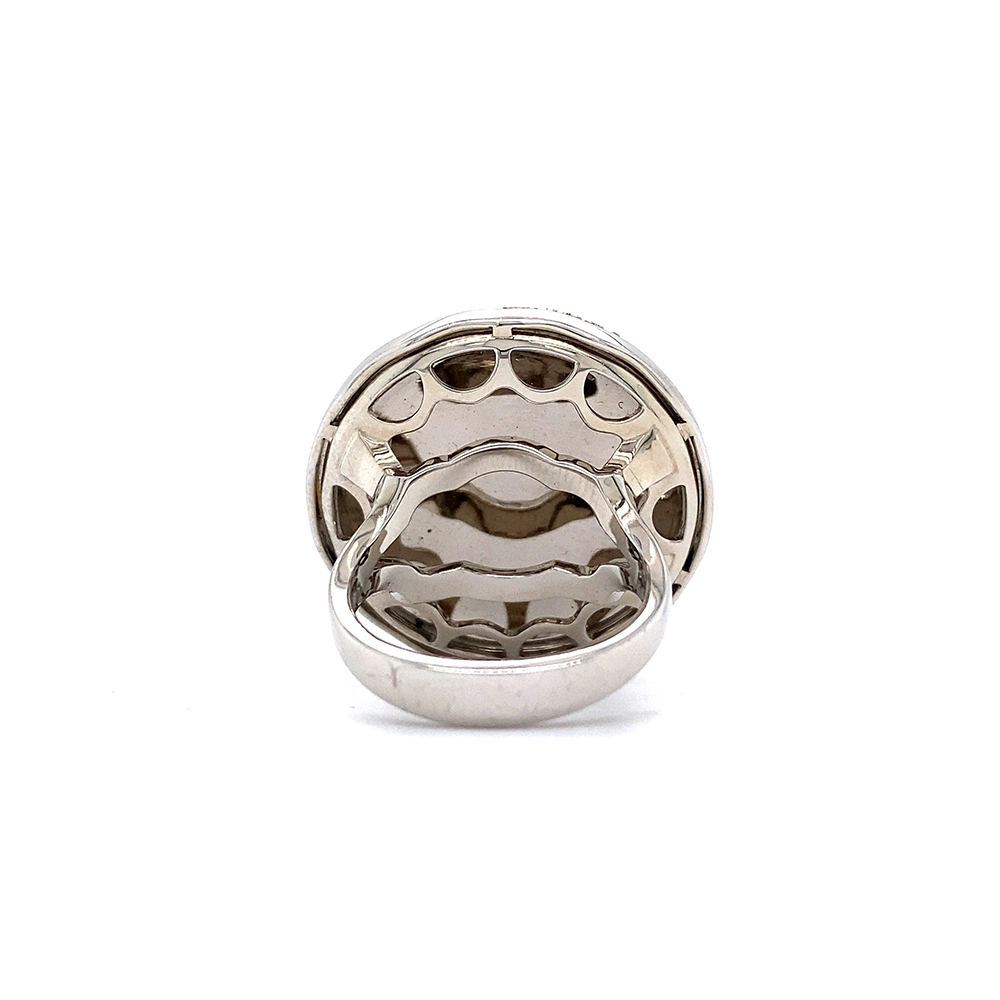 A ring made of white gold with a round head. The head is about the size of a large coin. Its surface shimmers in a strong golden-amber tone with a wave-like pattern. On top lies the shape of a large flower set with many small diamonds.