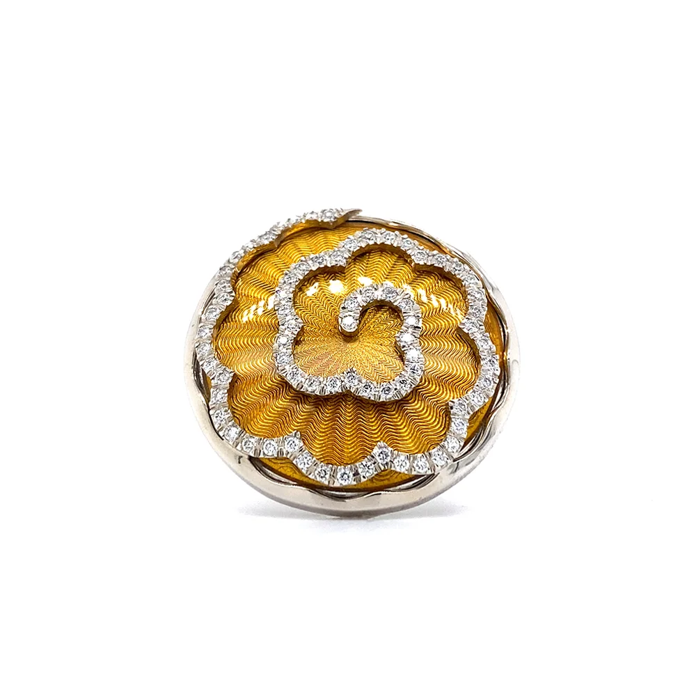 A ring made of white gold with a round head. The head is about the size of a large coin. Its surface shimmers in a strong golden-amber tone with a wave-like pattern. On top lies the shape of a large flower set with many small diamonds.