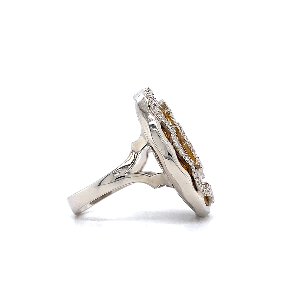 A ring made of white gold with a round head. The head is about the size of a large coin. Its surface shimmers in a strong golden-amber tone with a wave-like pattern. On top lies the shape of a large flower set with many small diamonds.