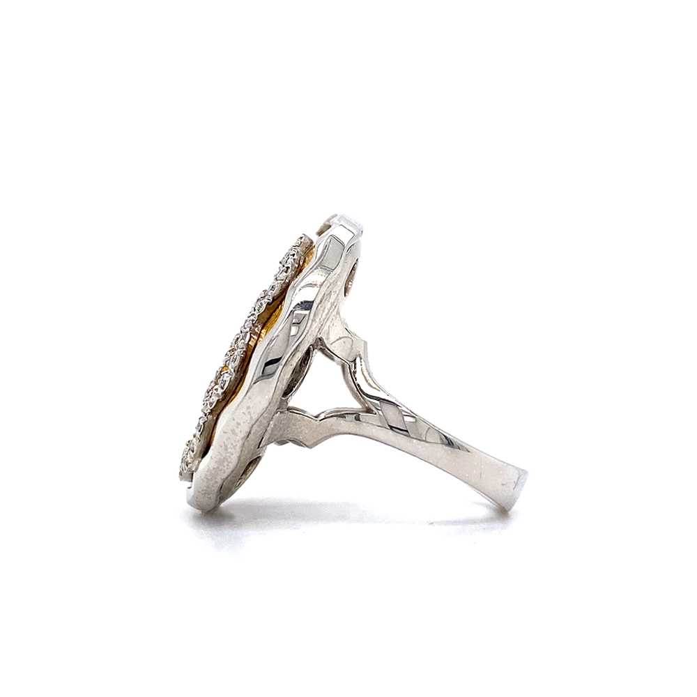A ring made of white gold with a round head. The head is about the size of a large coin. Its surface shimmers in a strong golden-amber tone with a wave-like pattern. On top lies the shape of a large flower set with many small diamonds.
