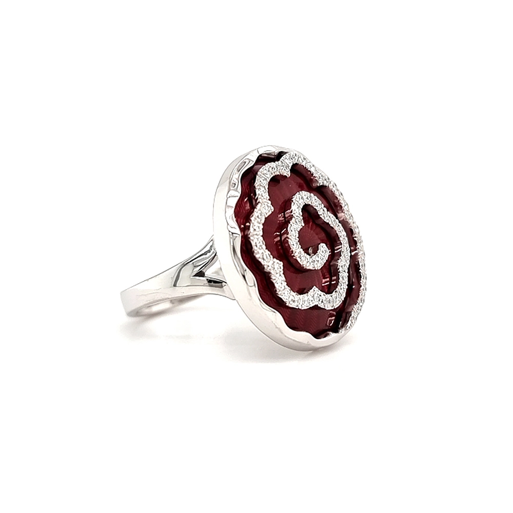 Ein Ring aus Weißgold mit einem runden Ringkopf. Dieser hat die Größe einer großen Münze. Die Oberfläche des Ringkopfes schimmert in einem kräftigen Rubinrot-Ton mit einem wellenartigen Muster. Darüber liegt die Form einer großen Blüte, besetzt mit vielen kleinen Diamanten.