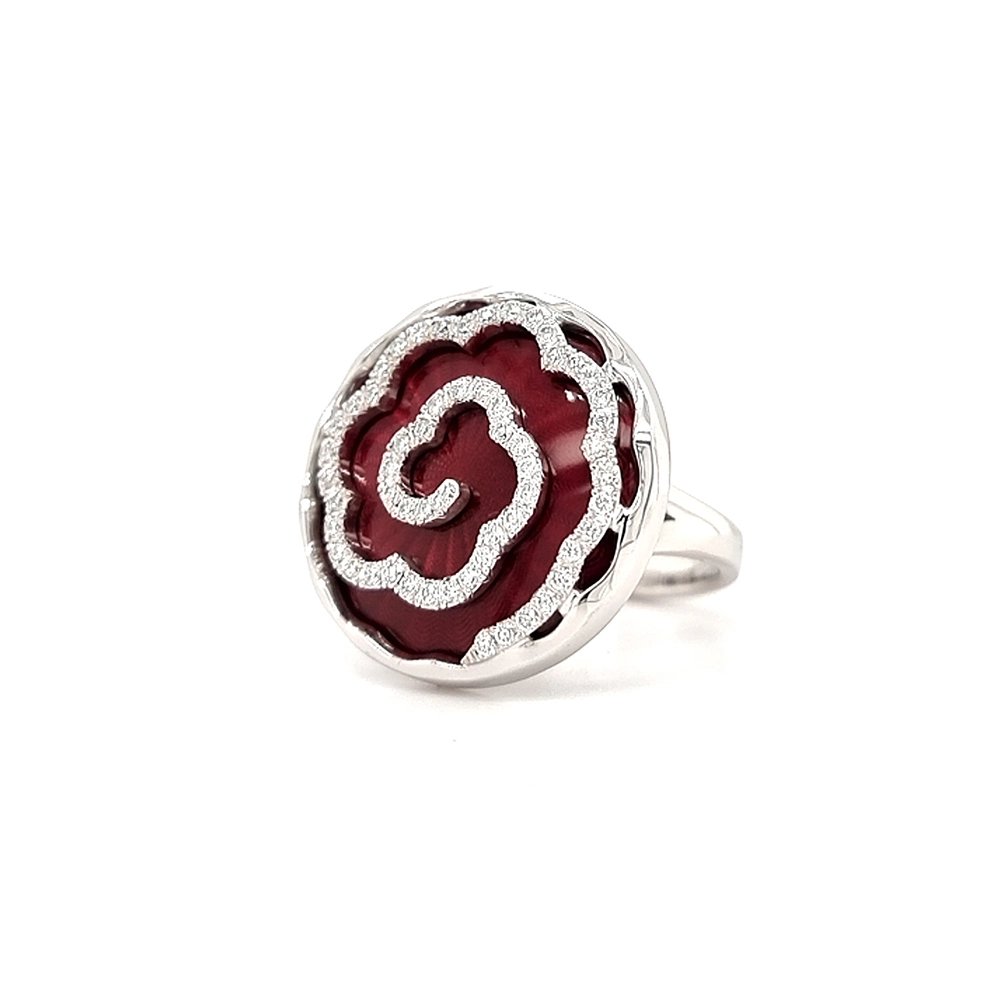 Ein Ring aus Weißgold mit einem runden Ringkopf. Dieser hat die Größe einer großen Münze. Die Oberfläche des Ringkopfes schimmert in einem kräftigen Rubinrot-Ton mit einem wellenartigen Muster. Darüber liegt die Form einer großen Blüte, besetzt mit vielen kleinen Diamanten.