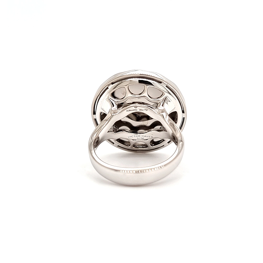 Ein Ring aus Weißgold mit einem runden Ringkopf. Dieser hat die Größe einer großen Münze. Die Oberfläche des Ringkopfes schimmert in einem kräftigen Rubinrot-Ton mit einem wellenartigen Muster. Darüber liegt die Form einer großen Blüte, besetzt mit vielen kleinen Diamanten.