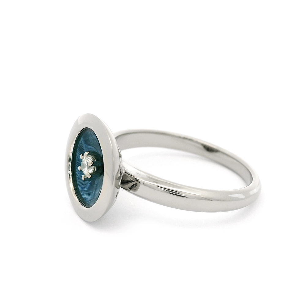 Ring aus Weißgold mit rundem Ringkopf. Der Ringkopf ist konkav gewölbt und mit mediumblauer transluzider Feueremaille versehen, unter der ein feines Windmühlen-Guilloché sichtbar ist. Im Zentrum der Emaillefläche sitzt ein einzelner kleiner Brillant als klarer Lichtpunkt. Die Gestaltung wirkt ruhig, geometrisch und ausgewogen.
